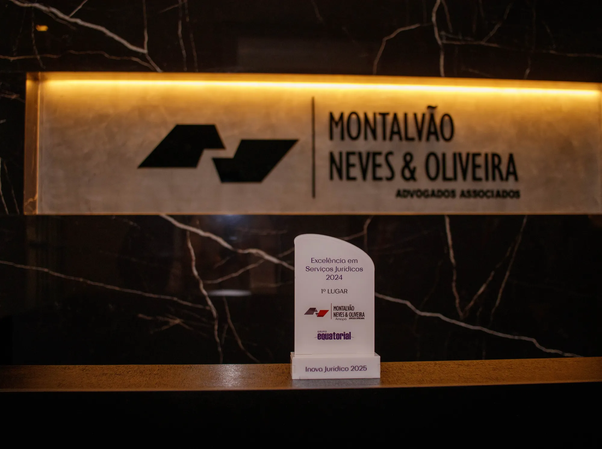 Recepção da banca em mármore preto retroiluminado, com o letreiro Montalvão Neves & Oliveira e o troféu de excelência em serviços jurídicos.